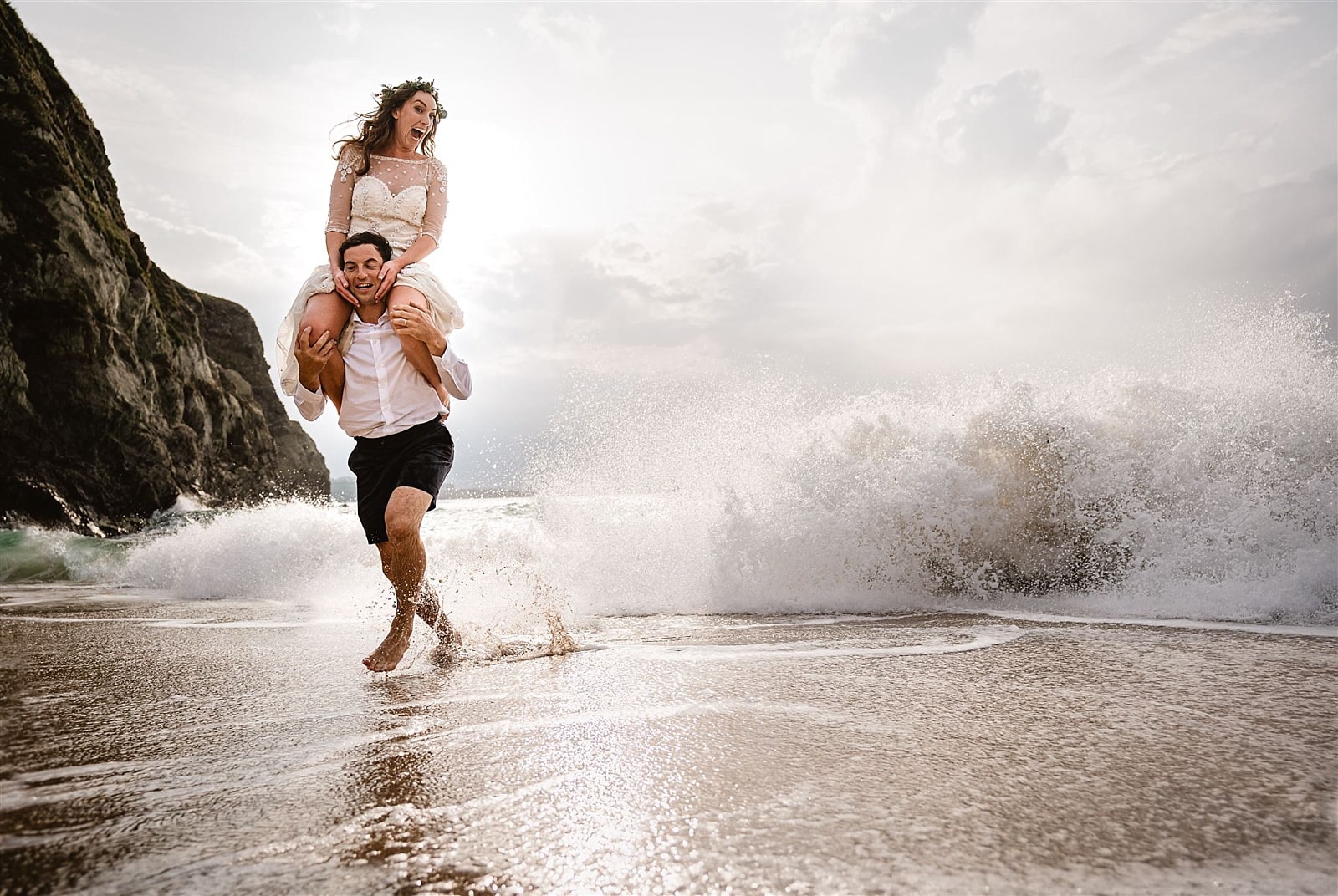 Lusty Glaze Wedding Photography: A newly wed couple run laughing from a crashing wave
