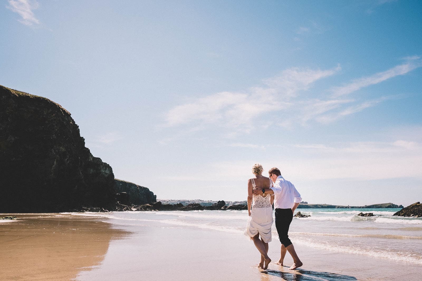 The wedding couple walking across Lusty Glaze beach in the sun.
