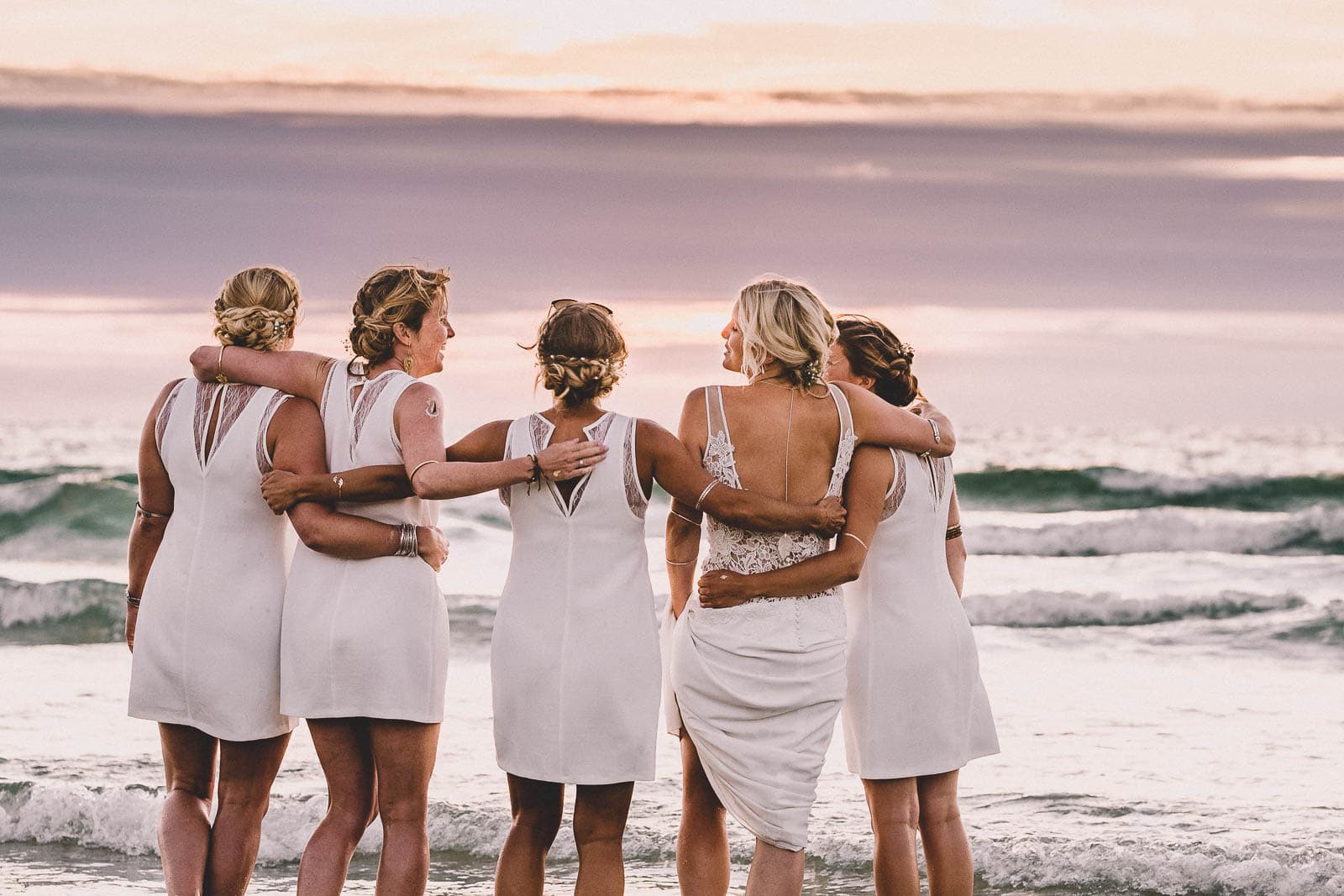 Lusty Glaze Wedding Photography. A bride and bridesmaids watch the sun setting on Lusty Glaze beach.