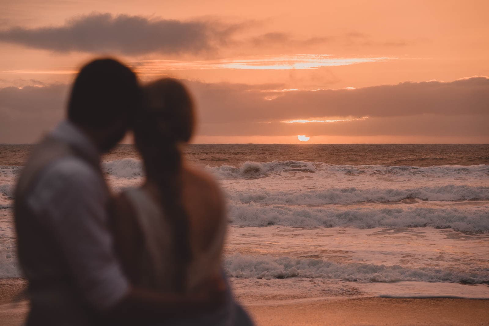 Lusty Glaze Wedding Photography. A stunning orange sunset at Lusty Glaze beach, with the bride and groom watching on.